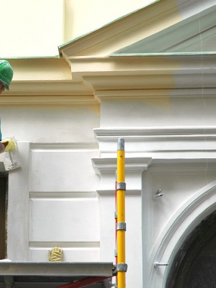 A construction worker in a green helmet and safety harness uses a paint roller on the exterior wall of a building, standing beside a scaffold.